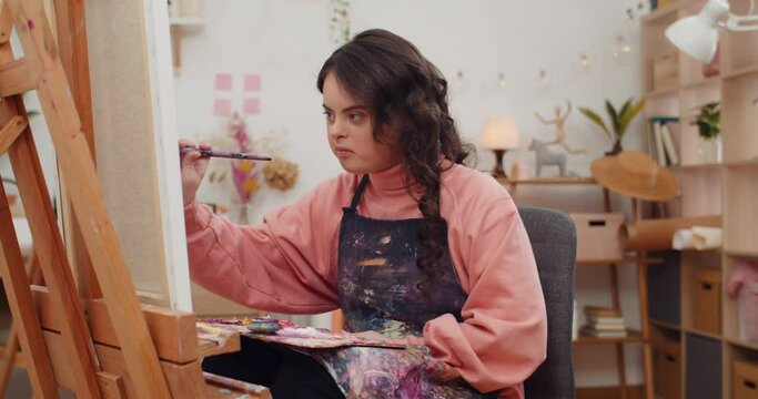 Pretty teenager girl wearing artist aprone soaking brush into paint. Adorable child with down syndrome holding art palette and painting while sitting in front of molbert in her room.