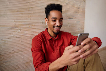 Glad young handsome short haired dark skinned curly guy holding mobile phone and smiling happily while reading messages, posing over beige interior in red shirt