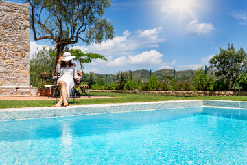 Obraz na płótnie Canvas A luxurios woman in white dress and hat sits by the pool and enjoys a hot summer day at her villa