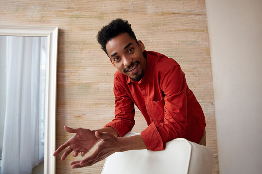 Glad Young Brown-eyed Dark Skinned Brunette Guy With Short Haircut Looking Positively At Camera While Leaning On Chair, Standing Over Home Interior In Red Shirt