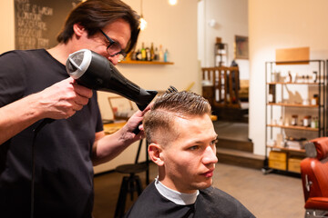 Barber dries the hair of man with hairdryer and brush