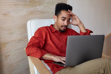 Concentrated young bearded dark skinned man with short haircut keeping raised hand on his head while checking email box on his laptop, isolated over home interior