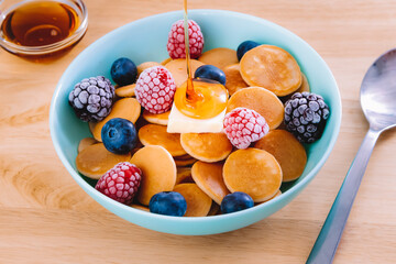 bowl of cereal pancakes with raspberries, blueberries and blackberries with butter and syrup being poured on next to a syrup container and a metal spoon on a wooden surface