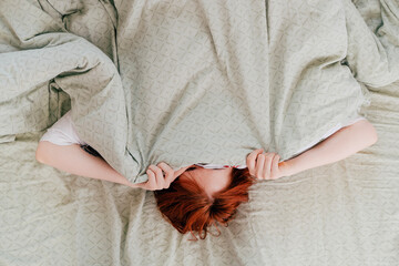 red-haired girl is hiding under the covers on the bed with soft green bedding
