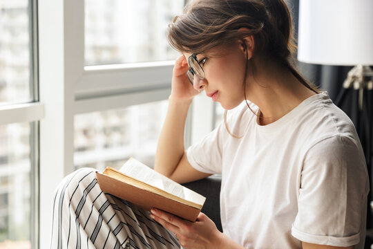 Photo of young thinking woman in eyeglasses reading book while sitting