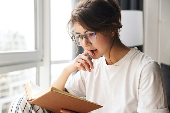 Photo of young sad woman in eyeglasses reading book while sitting