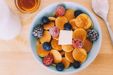 Top view of a blue bowl of cereal pancakes with berries and butter next to some syrup on a wooden table