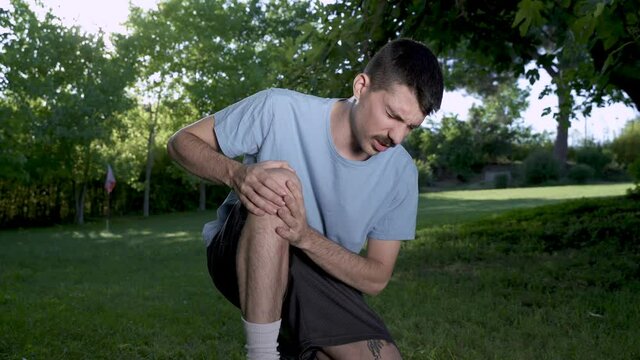 Male runner in pain on injury at the knee the leg on a park exercising or practicing for a marathon race, wearing sport clothes on sunny sunset sky. Spring time.