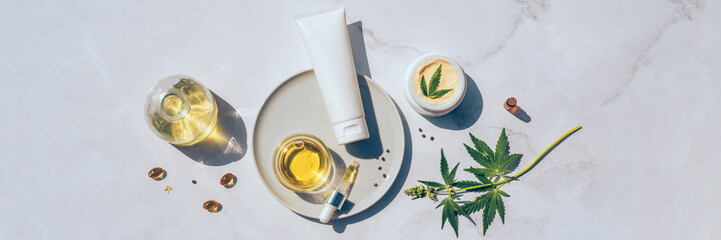 Tuba and jar of cream CBD oil, THC tincture and hemp leaves on marble background. Flat lay,...