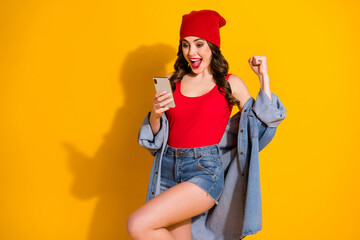 Positive youth student girl rejoice use smartphone celebrate win social media news impressed raise fists scream wear denim headwear red singlet isolated bright shine yellow color background