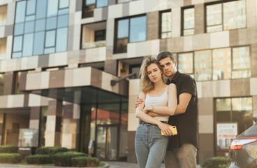 Young family stands on the street against the backdrop of a modern apartment building, hugging and looking at the camera, wearing stylish casual clothes. .