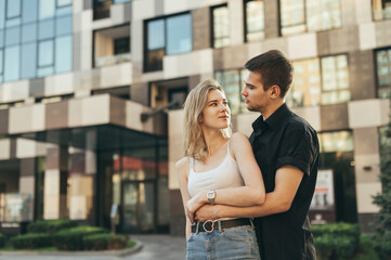 Beautiful young couple hugging on the street on the background of a modern building, the girl looks away. Guy hugged the girl on a walk on a warm summer day, she is happy. Love story