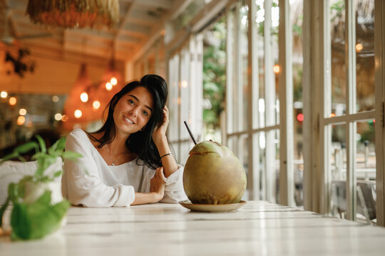 Focused optimistic attractive young  woman in summer clothes resting at table enjoying refreshing juice from coconut in cafeteria on blurred background