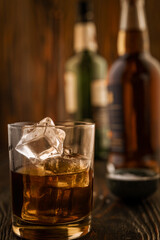 A glass of whiskey with ice, in the background are bottles on a wooden table of a bar counter, shallow depth of field, selective focus. The concept of alcoholic drinks in a roadside bar.