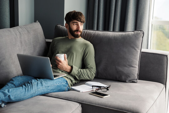 Image of man using laptop and drinking coffee while sitting on sofa