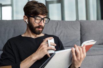 Image of focused man drinking coffee and reading magazine while sitting