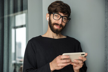 Image of young smiling man using wireless earphones and mobile phone