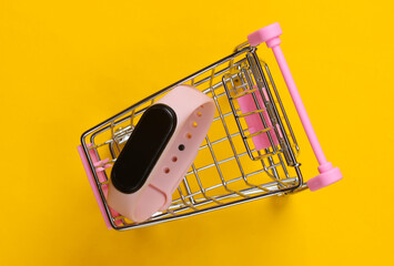 Mini shopping trolley with smart bracelet on yellow background. Modern gadgets