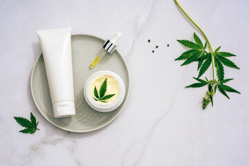 Tuba and jar of cream CBD oil, THC tincture and hemp leaves on marble background. Flat lay,...