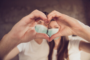 Closeup photo of cute young couple lady guy spend quarantine together stay home happy life make arms fingers heart figure faces inside sign sit sofa wear facial medical mask room indoors