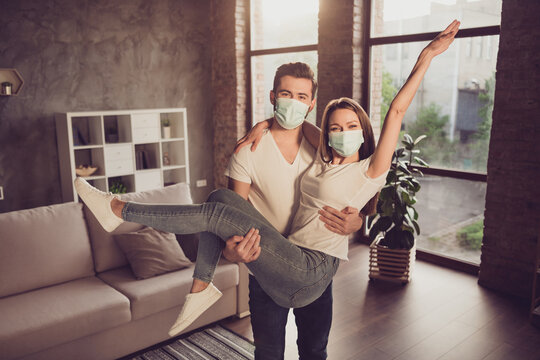 Photo of young couple guy hold arms excited lady quarantine together stay home hugging happy have fun just married moved new flat wear facial medical mask living room evening sunset indoors