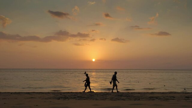 Silhouettes of adult people are walking in water edge on sunset tropical beach with dark waves splashes and jet ski in sea