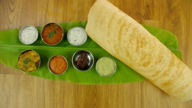 Zoom out shot of South Indian dish masala dosa served over a fresh banana leaf. Top view shot of South Indian breakfast meal dosa with bowls of sambhar  fried red chilly and colorful chutneys