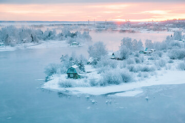 Cold orange dawn with gentle fog over wooden dacha houses on Angara River island, snowy trees and frozen water, Irkutsk, Siberia