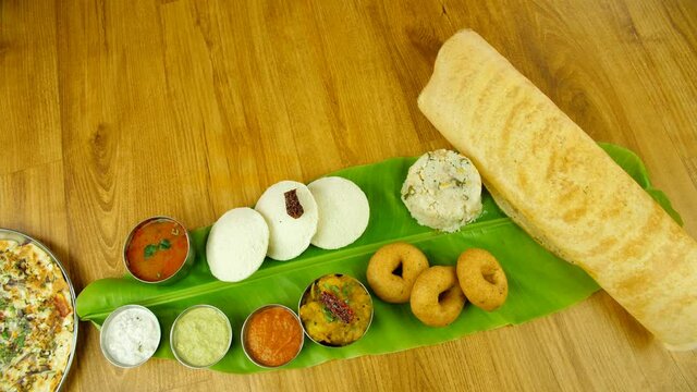 Popular traditional South Indian platter served over a banana leaf on a wooden table. Group of South Indian food like Dosa  Idli  Upma  Uttapam  Vada served with potato masala  sambhar  and chutneys