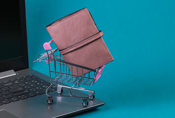 Mini supermarket trolley with wallet on laptop keyboard. Blue studio background. Online shopping