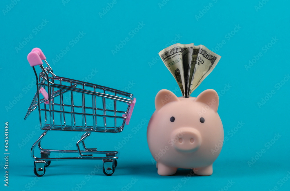 Wall mural shopping concept. piggy bank with dollar bil and miini supermarket trolley. blue studio background.