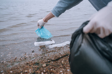female volunteer removes debris from used medical masks and gloves on the beach and in the water