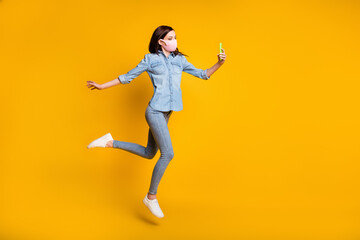 Fototapeta na wymiar Full length photo of beautiful girl jump use smartphone make selfie video call connection technology wear medical mask denim jeans shirt isolated over yellow color background