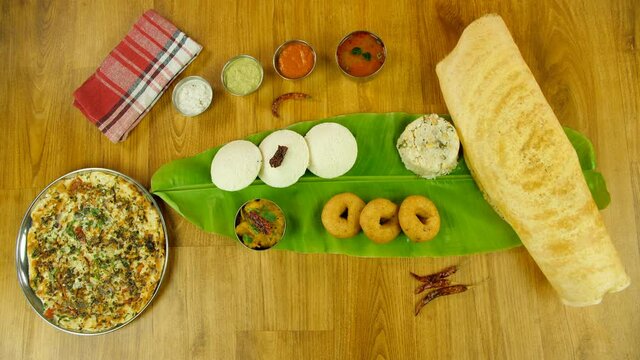 Woman hands serving beautifully plated vegetable Uttapam on a wooden platform. Group of South Indian food like Dosa  Idli  Upma  Uttapam  Vada served with potato masala  sambhar  and chutneys