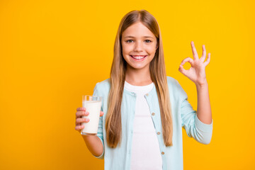 Photo of beautiful little blond lady hold arm fresh milk glass show okey symbol advising drink dairy products every morning healthy wear casual blue shirt isolated yellow color background