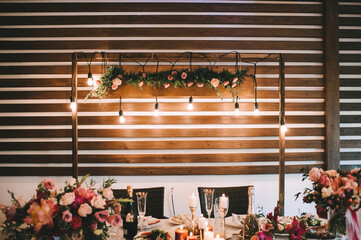 Rustic, wooden background, roof, garland of light bulbs, interior. The table is covered with a white tablecloth, served, compositions of flowers and greenery. Plates, cutlery, napkin, blue glasses.
