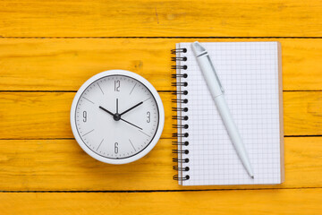 Fototapeta na wymiar White clock and notebook on yellow wooden background. Minimalistic studio shot. Top view