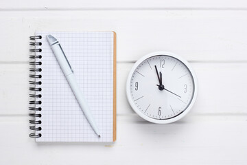 Fototapeta na wymiar White clock and notebook on white wooden background. Minimalistic studio shot. Top view
