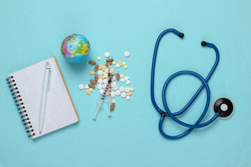 Syringe and globe, pills and notebook, stethoscope on blue background. Global vaccination, treatment. Covid-19 pandemic. Top view