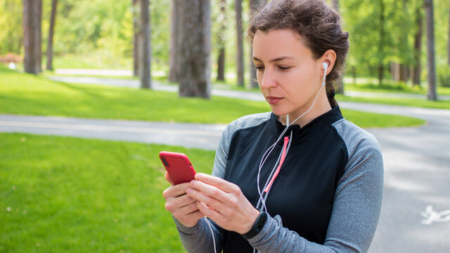 Young concentrated sporty woman in headphones holding smartphone and looking at screen,sharing running data on social media,listening music on mobile phone after jogging,training,work out 