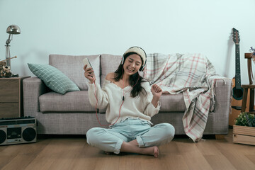 Fototapeta premium happy young woman in headphones listening music sitting comfortably on floor with sweater and bobble hat. copy space. joyful lady dancing moving body with melody in headphones from cellphone online