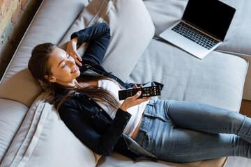 overhead view of happy woman holding remote controller near laptop with blank screen on sofa