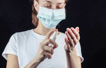 Woman in a median face mask disinfects hands with an antiseptic on a black background. Hygiene in...