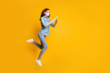 Full size photo of beautiful girl jump use smartphone read social covid network news wear medical mask denim jeans shirt isolated over bright shine color background