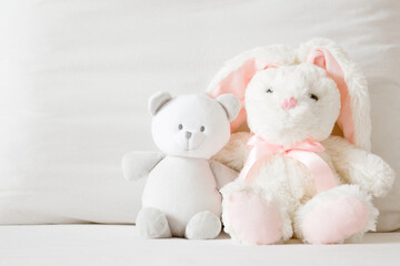 Fototapeta na wymiar Smiling white teddy bear and fluffy bunny sitting on bed at pillow. Togetherness and friendship concept. Kids best friends. Front view. Closeup.