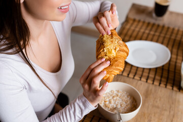 cropped view of cheerful girl holding croissant near bowl with corn flakes