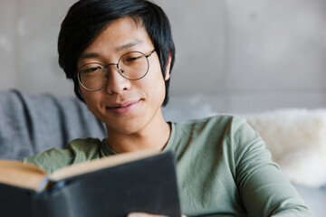 Image of young asian man wearing eyeglasses reading book in apartment - Powered by Adobe