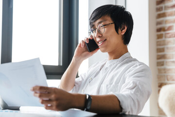 Image of asian man using cellphone and holding paper documents in office