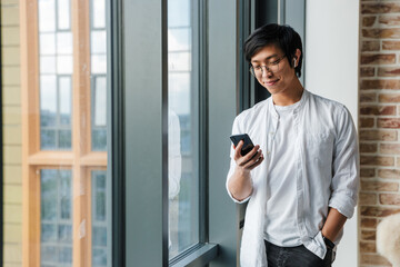 Image of young asian man wearing earphones using cellphone in office
