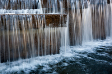 beautiful cascading artificial waterfall from stone and concrete blocks on the city water river channel with blurry streams and a bubbling foamy surface of the water